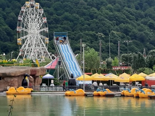 Amusement park with roller coasters, pedal boats & water slides against a mountain backdrop. Fun for kids and adults.